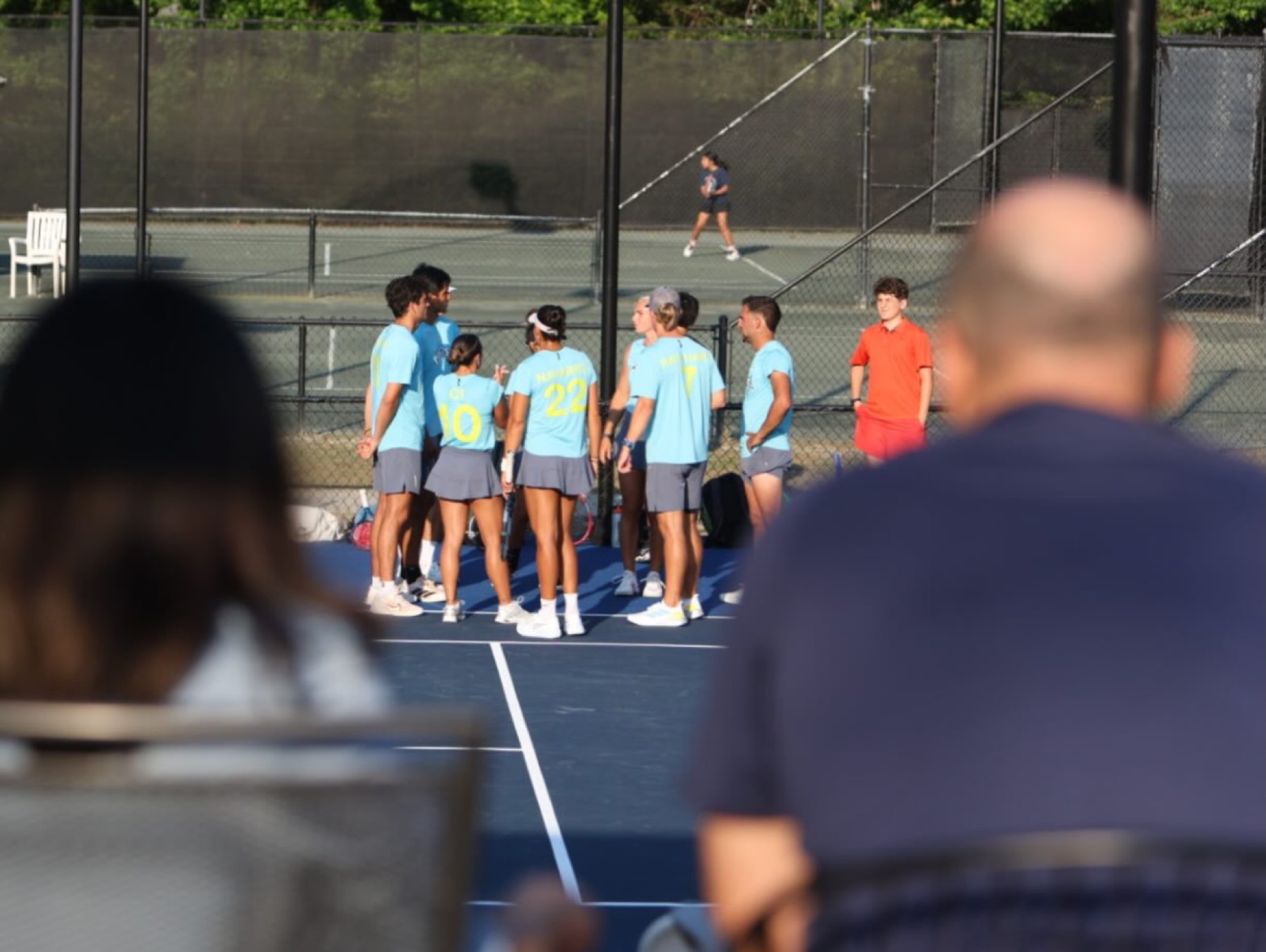 A team huddle on a club court seen over the shoulders of club members who have stopped to watch courtside.